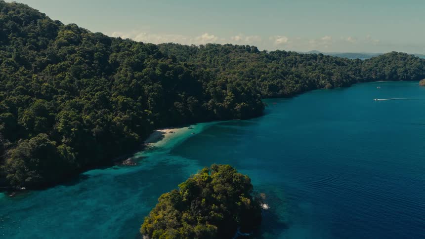 A lush green island surrounded by vibrant turquoise and deep blue ocean water and a small boat moves in the distance
