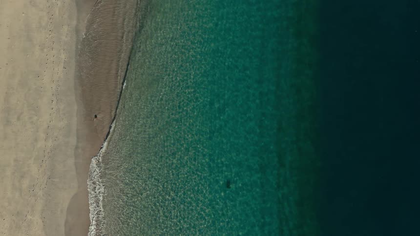 An aerial shot captures a sandy beach meeting clear turquoise water that transitions into a deep navy blue small waves gently lap onto the shore while the camera remains fixed in a topdown perspective