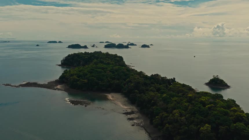 An aerial shot captures a lush green tropical island surrounded by calm turquoise waters under a cloudy sky the camera glides smoothly over the coastline revealing smaller neighboring islands scattered across the distant horizon