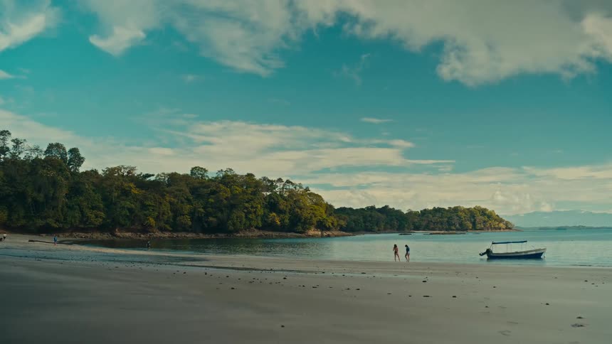 Two people walk slowly along the shoreline of a wide sandy beach while a small boat floats nearby in the calm blue watera dense green forest lines the edge of the coast under a bright sky filled with soft white clouds