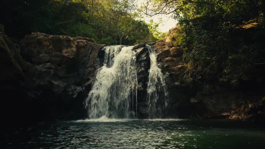Waterfall flows over rocks on a mountain cliff in the forest as a young man swings on a rope and drops into the water