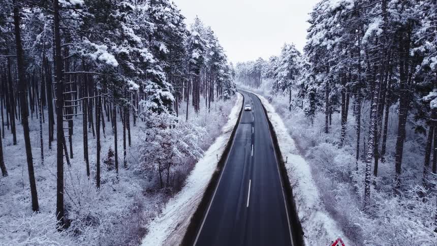 An aerial view shows cars driving along a dark asphalt road that cuts through a dense forest where every pine tree and the ground are covered in thick white snow under a grey winter sky