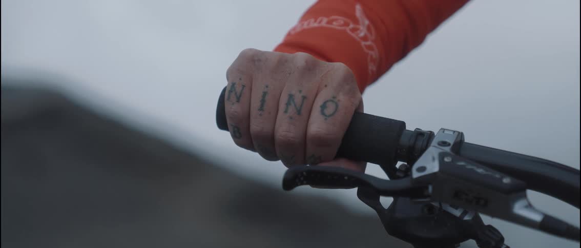 A tattooed hand reaches out and firmly grasps the black rubber grip of a mountain bike handlebar against a blurry outdoor backdrop