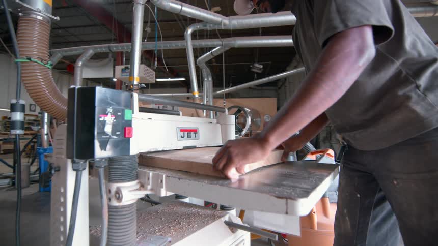 A carpenter wearing a tan cap and glasses works in a workshop where he lifts a thick piece of light wood and carefully guides it through a power planer machine to smooth the surface of the timber