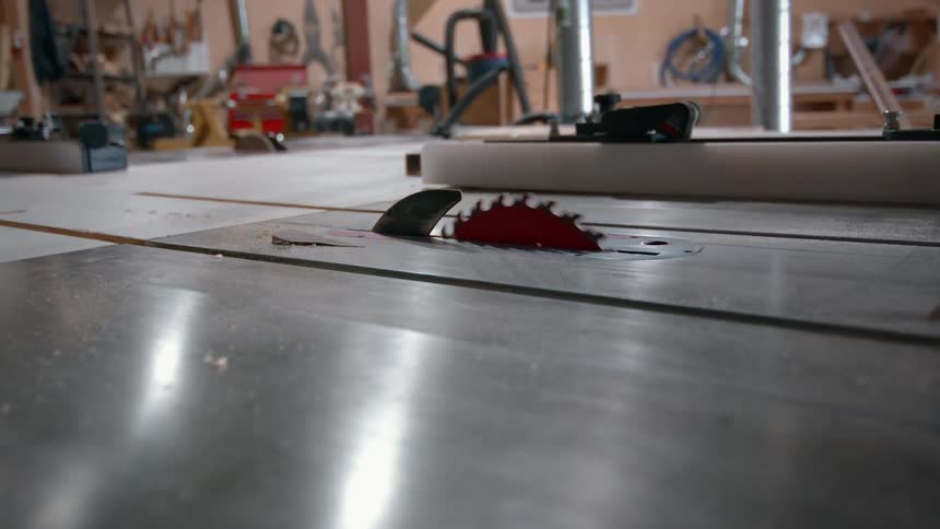 A red circular saw blade rises from a metal table and begins to spin in a professional woodworking workshop with various tools and equipment visible in the blurred background