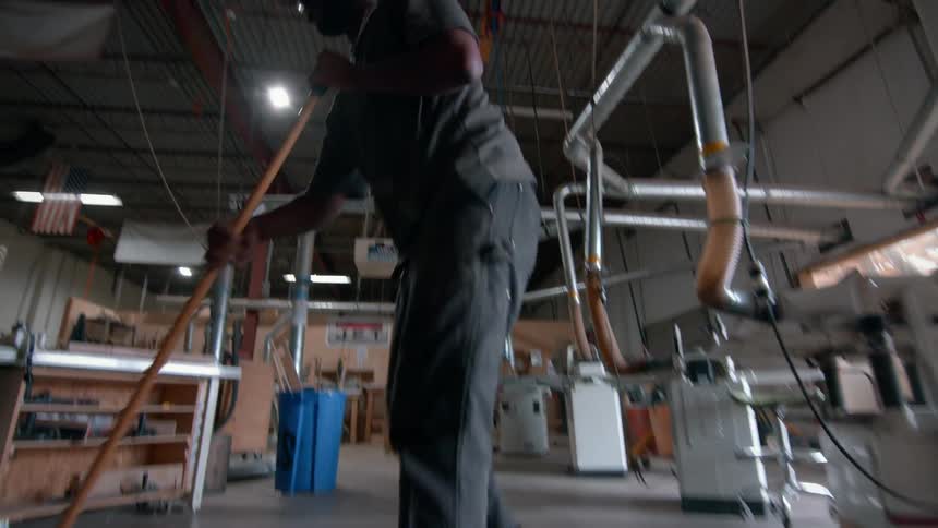 Man sweeps workshop floor with machines and ventilation ducts in the background