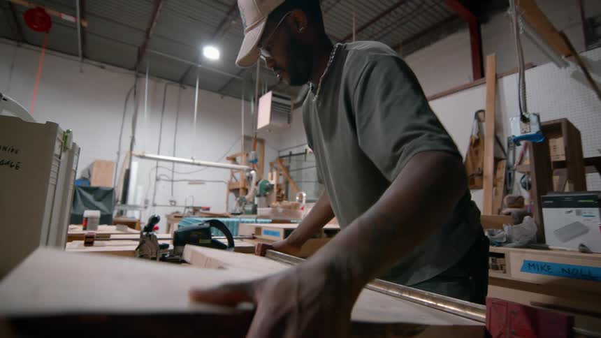 A man lifts a large wooden slab from a workbench in a woodworking shop