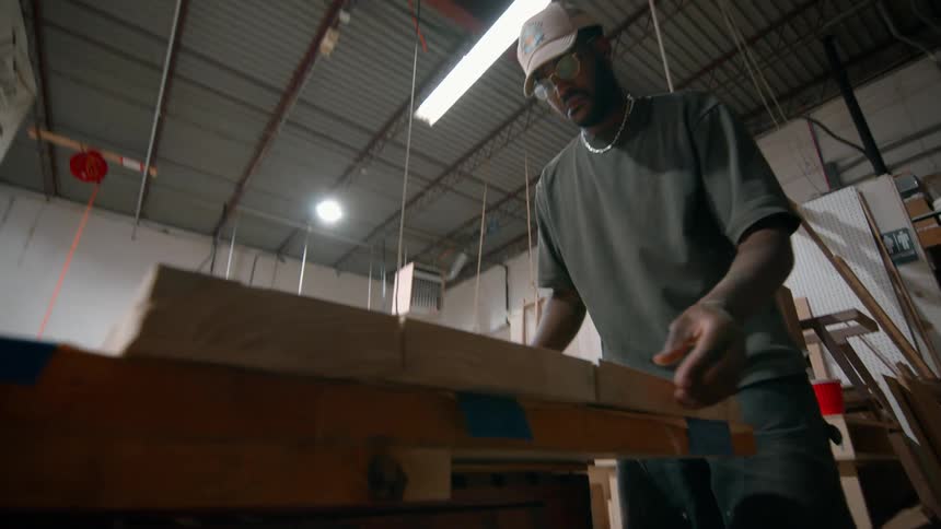 A male carpenter works inside a workshop assembling pieces of wood while wearing a hat and a necklace