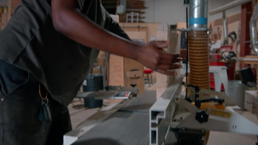 A carpenter wearing a tan cap and glasses works in a workshop where he lifts a thick piece of light wood and carefully guides it through a power jointer machine to smooth the edge of the lumber