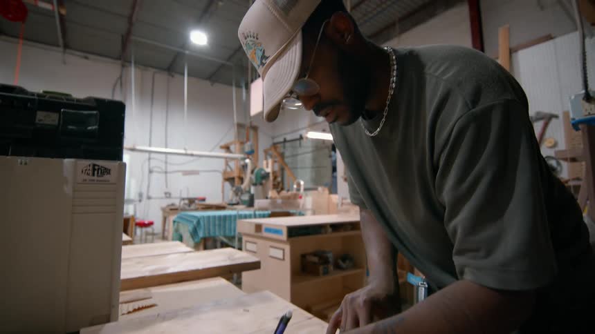 A carpenter wearing glasses a hat and a necklace marks a wooden piece on a workbench with a pen