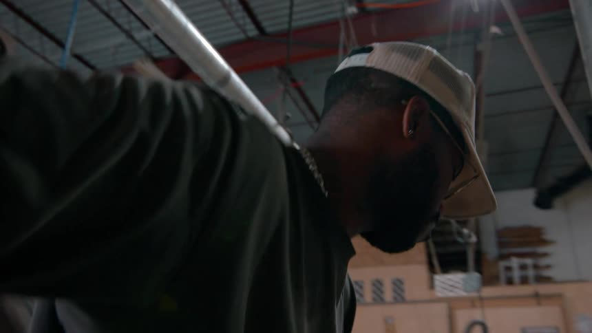 A carpenter wearing a tan cap and a dark shirt uses a jointer machine to plane the edge of a large wooden board in a professional workshop with industrial ventilation pipes and tools