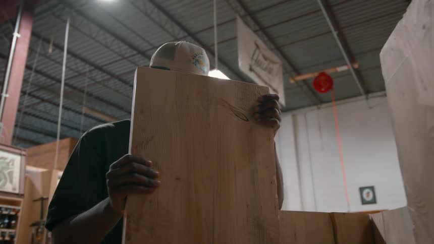 Carpenter standing in a workshop holds up a wooden board on the table