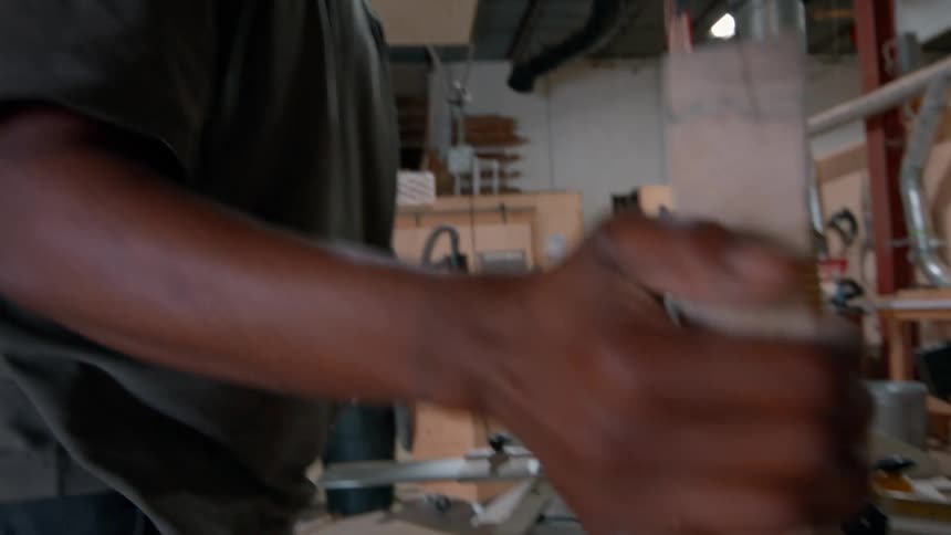 A male a focused woodworker with glasses and a cap carefully feeds a large live edge wooden slab through a jointer machine inside a large industrial workshop filled with ventilation pipes and specialized carpentry equipment