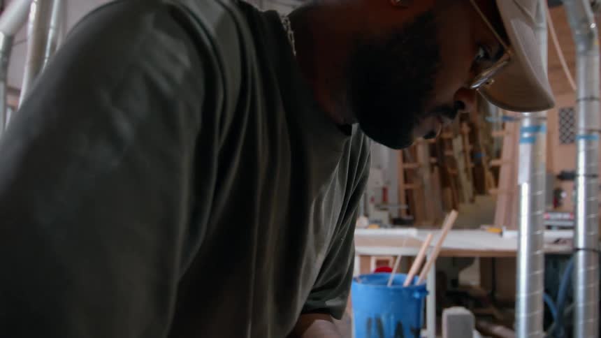 A man wearing a baseball cap and glasses is shown working intently in a woodworking shop he is focused on a piece of wood in the foreground while surrounded by various tools lumber and ventilation pipes in the workshop background