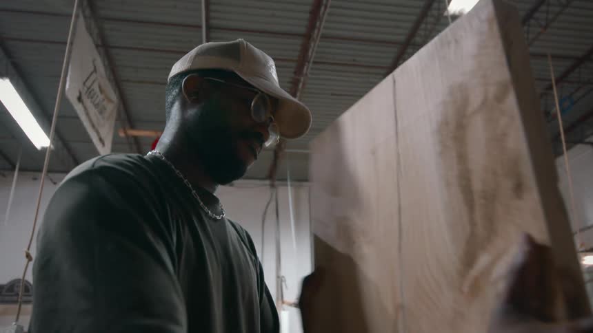 A man with glasses and a cap stands in a carpentry workshop carefully inspecting a large piece of raw timber
