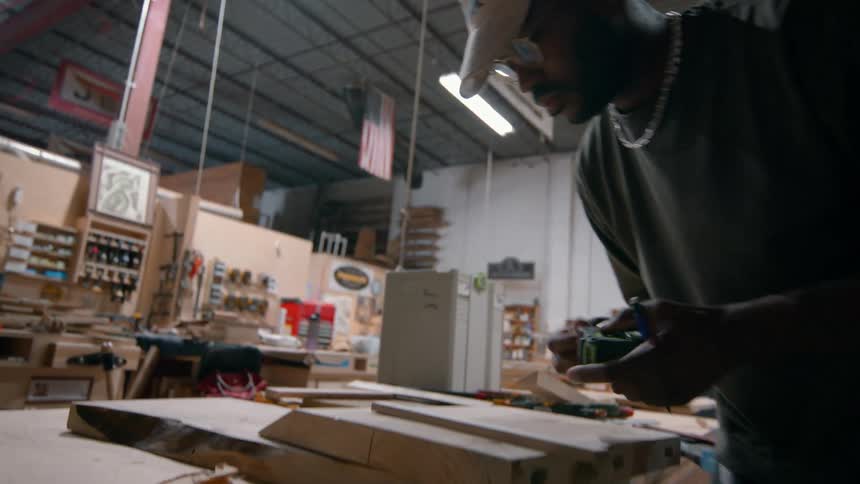 A male woodworker wearing a cap and silver chain uses a pencil and measuring tape to mark a light wooden plank on his workbench while surrounded by tools and various lumber materials in a large industrial carpentry workshop