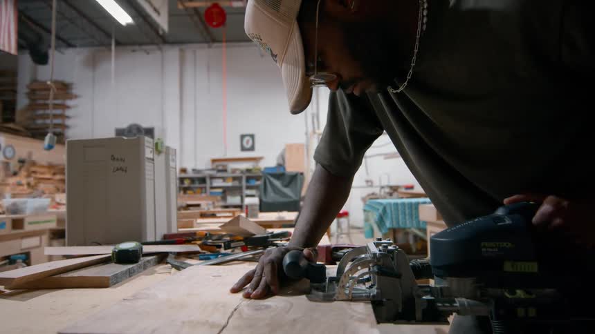 Man uses power tool to cut wood on workbench in workshop with tools scattered around