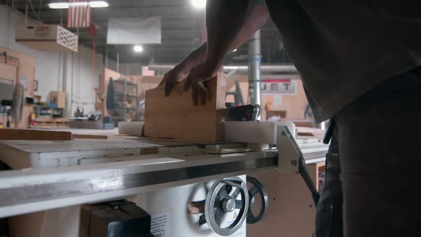A woodworker inspects a piece of lumber before carefully feeding it through a table saw to make a precise cut once the cut is complete he removes the wood and shuts off the machine in a large workshop setting