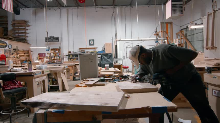 A man wearing a grey shirt and a white baseball cap uses a power tool to sand a large piece of wood on a workbench inside a busy carpentry workshop filled with tools and lumber