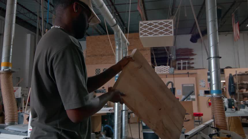 Man in baseball cap inspecting wood board before placing it on jointer machine inside workshop