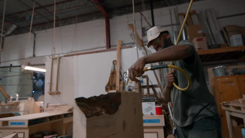 Carpenter standing in his workshop pulls an air hose from the ceiling and sprays air over the wooden board