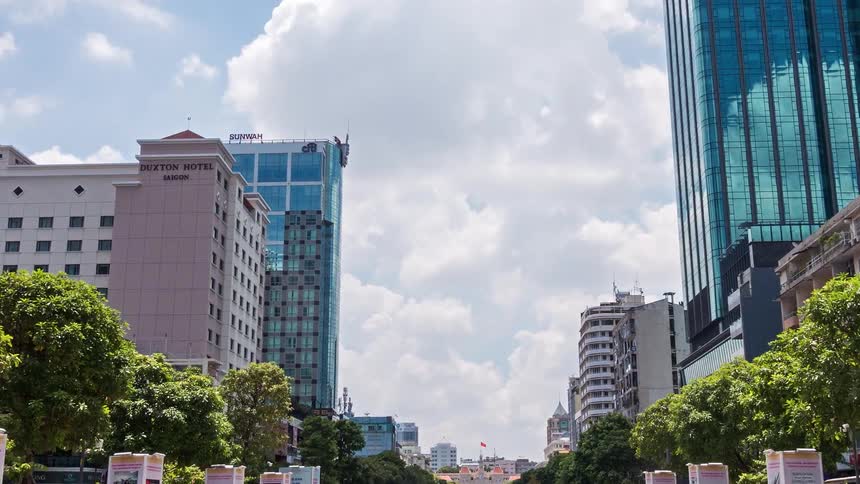 A wide pedestrian square stretches toward distant city buildings under a bright cloudy sky while rows of display posters and green trees line the walkway near tall glass skyscrapers