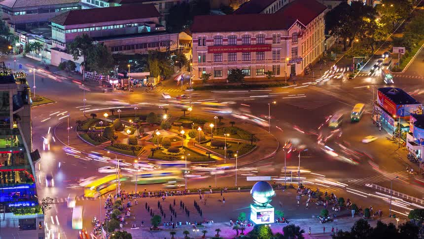 Light trails from vehicles moving around a busy city roundabout and adjacent square where people gather near a globe monument surrounded by illuminated multi story buildings and dense urban infrastructure