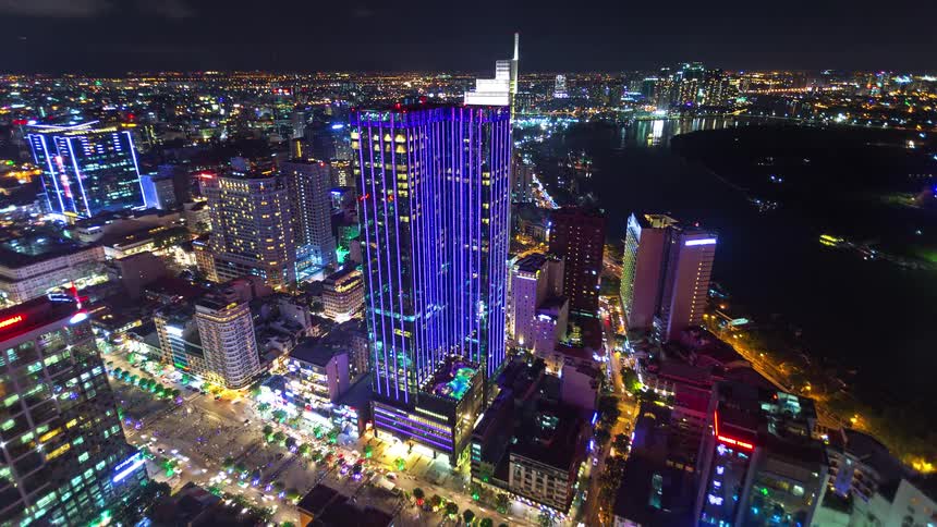 Lights glow on downtown buildings and flash on a tower as traffic moves on the streets of a metropolitan city on the coast and boats sail on the water