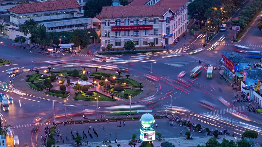 A high angle time lapse view shows many cars and motorbikes with bright lights moving quickly around a circular green park in a busy city intersection during the transition from dusk to evening