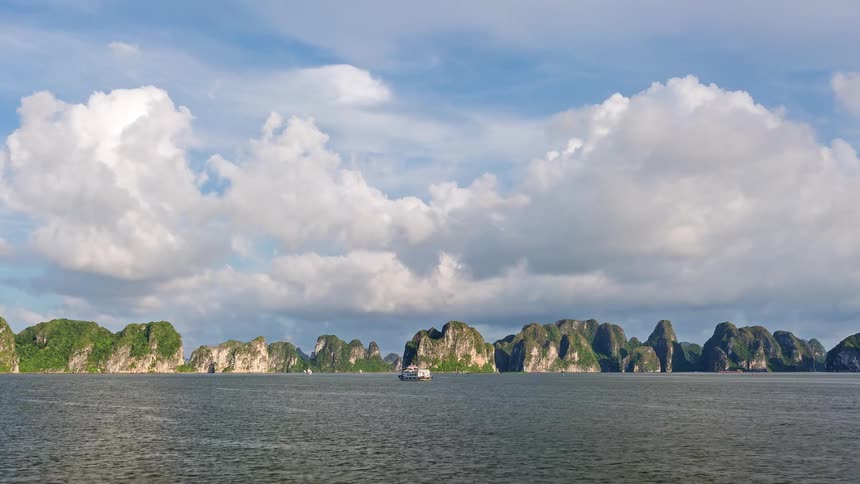 Series of boats moves across a lake at midday with a mountain range and rocky landscape lining the background