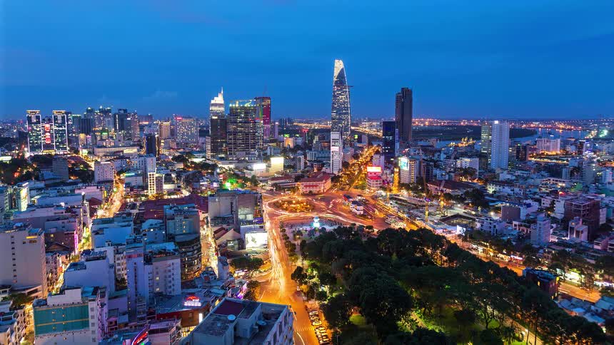 A high angle time lapse view shows many cars and motorbikes with bright lights moving quickly around a circular green park in a busy city intersection during the transition from dusk to evening