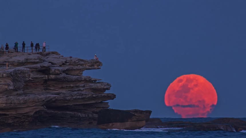 A massive orange moon rises from the horizon behind a jagged cliff where several people stand on an observation deck and one person sits near the edge while waves crash against the rocks under a deep blue twilight sky