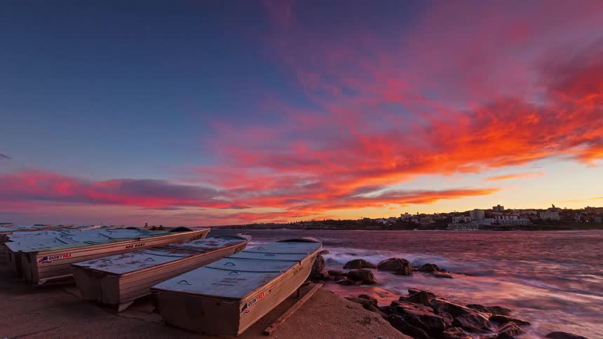 A timelapse of a coastal sunset where the sky transitions from bright orange to a deep fiery redin the foreground several small fishing boats are parked on a concrete ramp next to a rocky shoreline with crashing waves