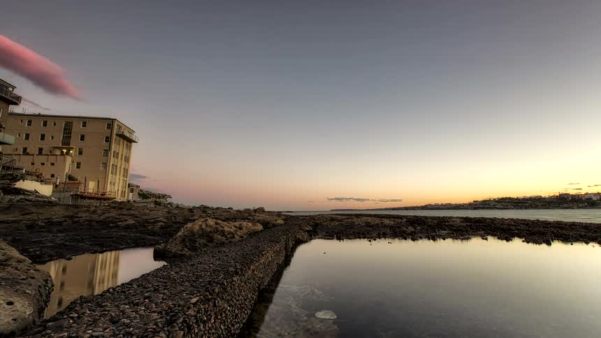 A stone walkway separating calm rock pools from the ocean under a sunset sky while a large tan building stands on the rocky coast