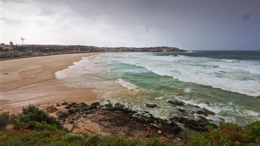Foaming green waves crash against a rocky shore and a wide sandy beach under a dark cloudy sky with a coastal town visible in the distance