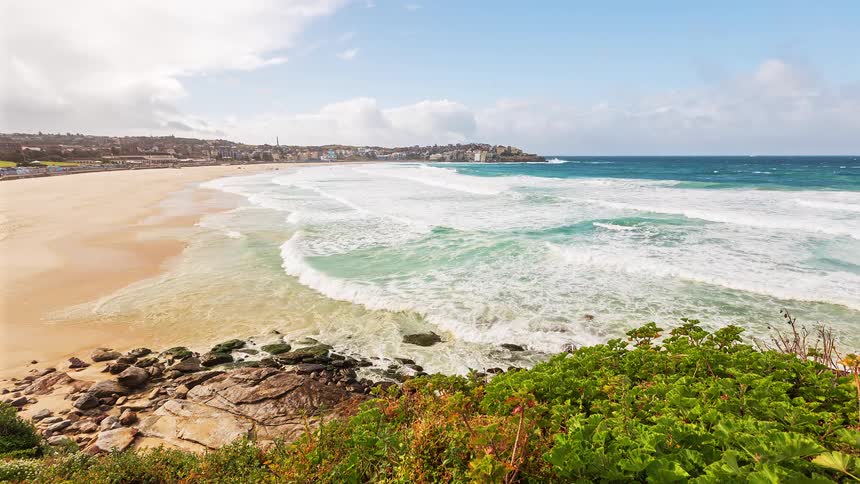 Wide beach with breaking waves rocks in the foreground and green vegetation by the seaside