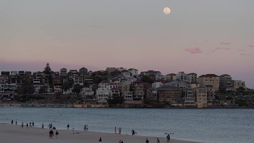 A beach filled with people with a city skyline across the water as the day fades into evening and the sun sets