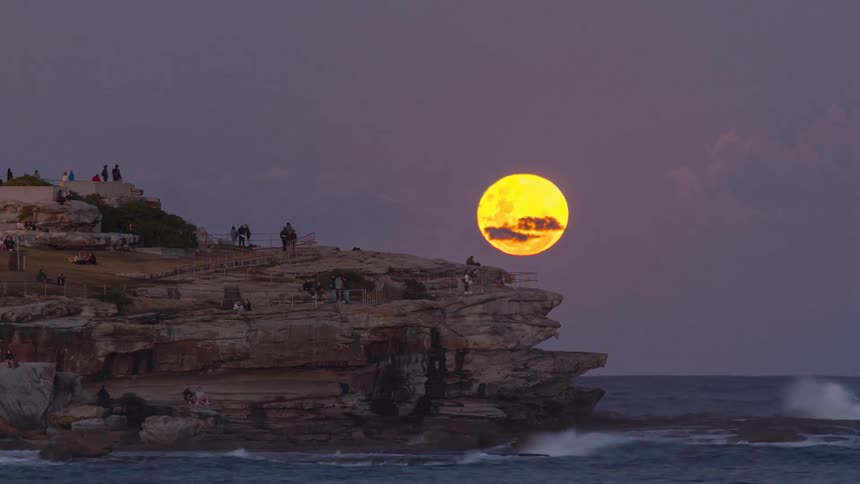 Full moon rises on the horizon behind rocky seaside formation at dusk