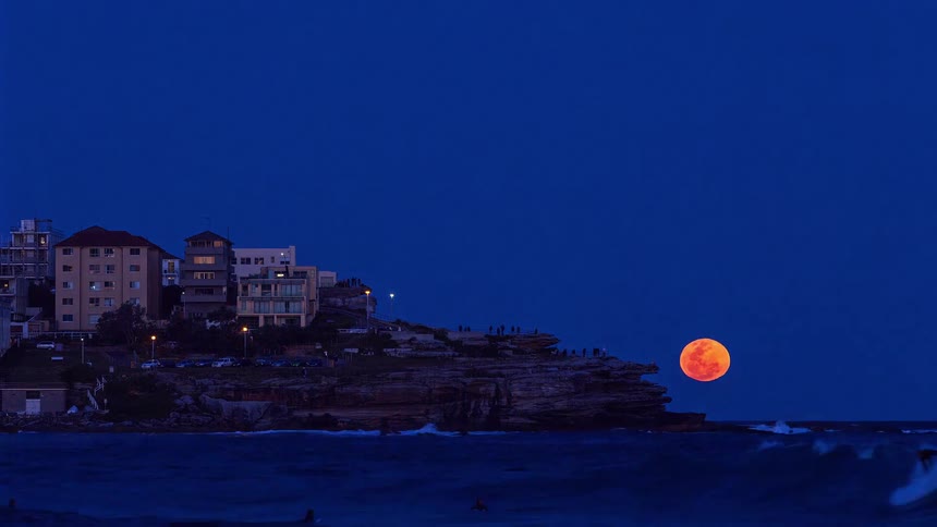 Buildings in a neighborhood on the coast with people standing on the cliff overlooking the water as the full moon rises in a clear sky