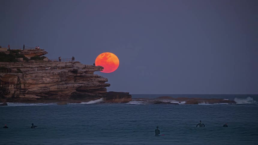 People standing on a cliff overlooking other people in the water as the full moon rises on the horizon