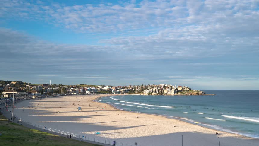 A wide sandy beach curves along the ocean where gentle waves roll toward the shore under a blue sky with soft white clouds and a coastal town visible in the distance