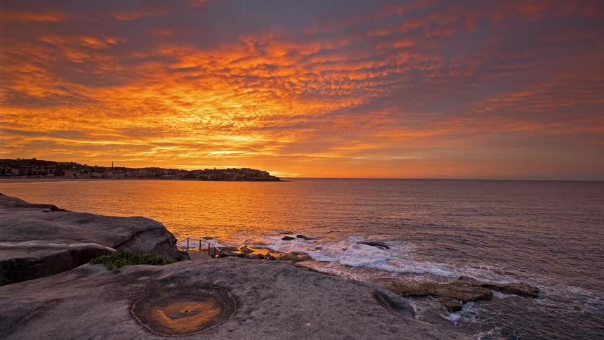 A fiery sunrise paints the morning clouds in deep shades of orange and red over a calm ocean baywaves gently wash against the rocky shoreline in the foreground as the sun begins to peek above a distant coastal town