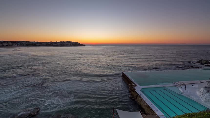The sun rises over the horizon at an ocean pool as waves crash against the concrete walls and theres a coastal town on the horizon
