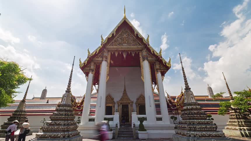 This timelapse captures a grand thai temple with ornate golden gables and tall spires under a bright cloudstreaked skya steady stream of visitors moves in and out of the entrance as soft shadows shift across the white temple walls throughout the day