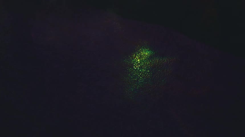 A dark textured surface illuminated by shimmering prismatic light that shifts between golden and green hues the play of light creates a fluid abstract effect as the bright reflections move across the shadowed background