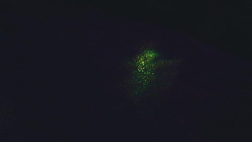 A dark textured surface illuminated by shimmering prismatic light that shifts between golden and green hues the play of light creates a fluid abstract effect as the bright reflections move across the shadowed background