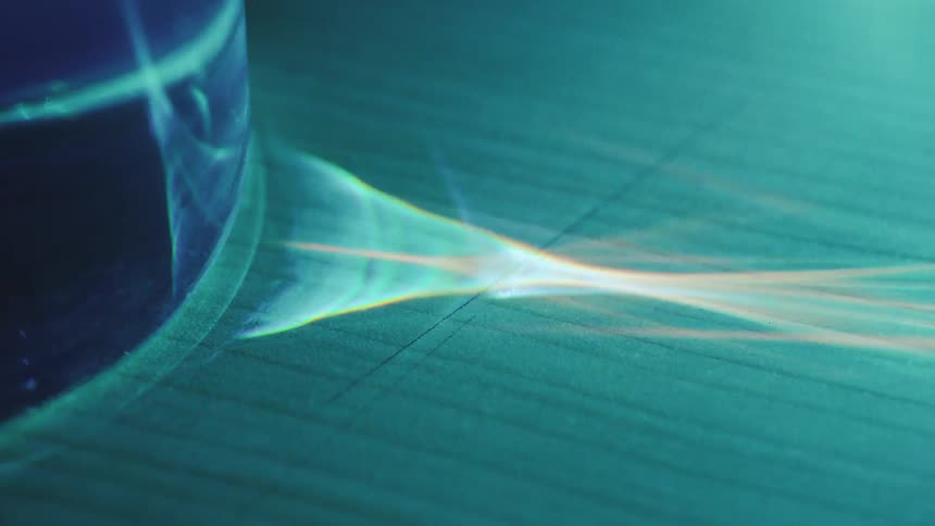 A close view shows bright blue light passing through a glass object and casting a sharp glowing beam with orange and yellow refractions across a dark textured surface
