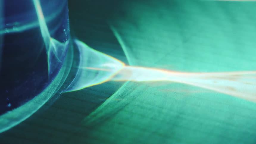 A close view shows bright blue light passing through a glass object and casting a sharp glowing beam with orange and yellow refractions across a dark textured surface
