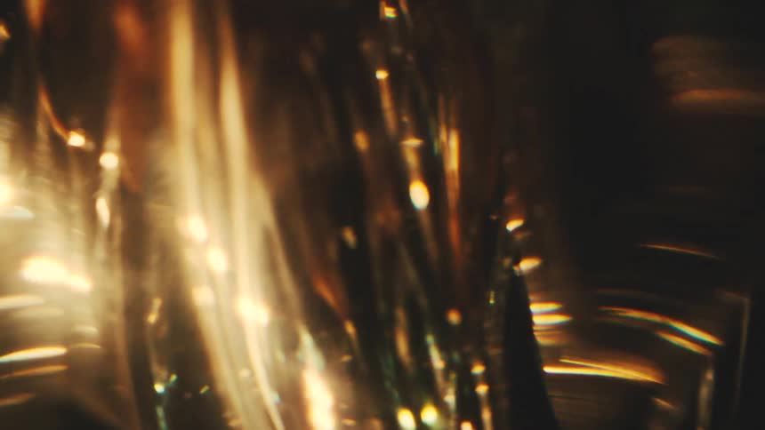 A close view shows warm golden light reflecting and shimmering through a textured glass surface against a dark background creating abstract shapes and bright flares