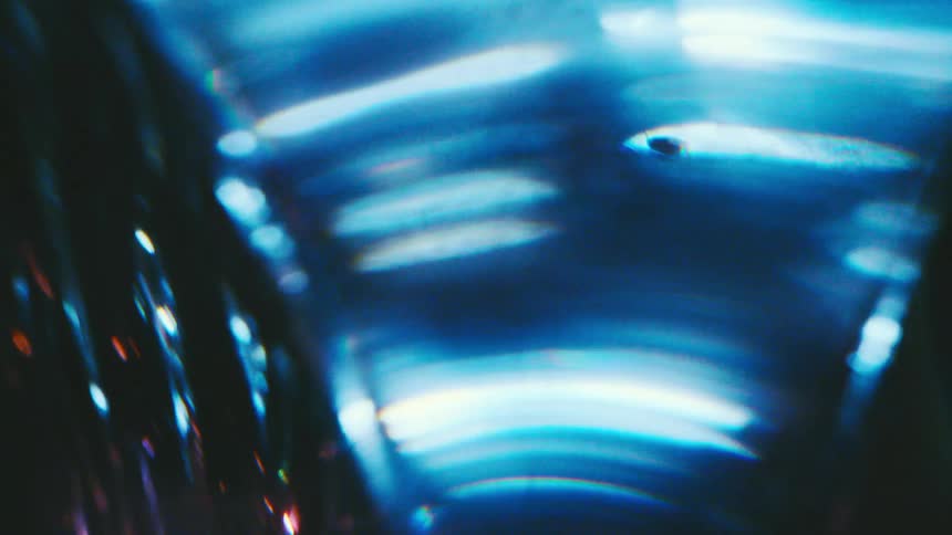 A close view shows bright blue and white light reflecting through textured glass with soft purple and orange glares moving across a dark background